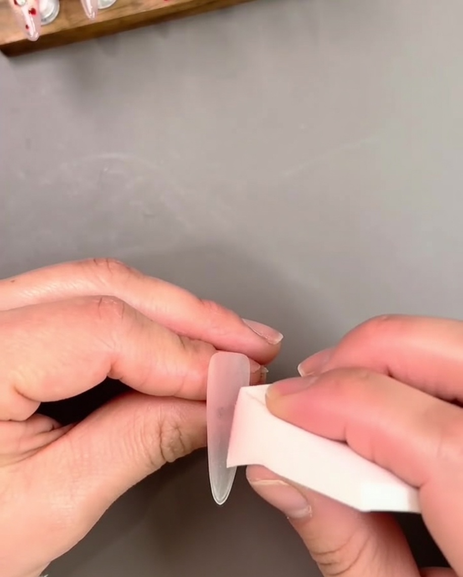 Una mano che sostiene unestensione per unghie mentre applica uno smalto gel rosa trasparente con un pennello.