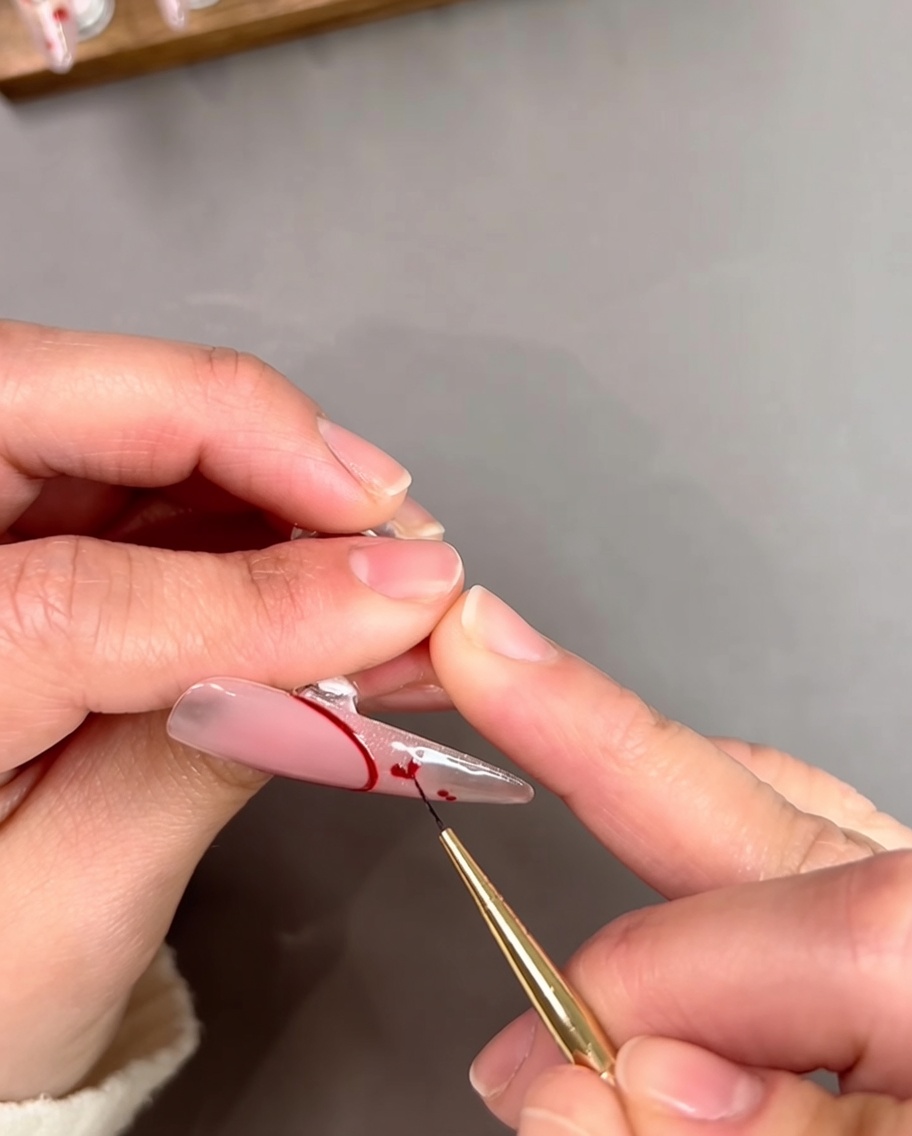 Un primo piano di una nail artist che usa un pennello sottile per collegare due piccoli punti rossi in una forma a cuore su una tip trasparente.