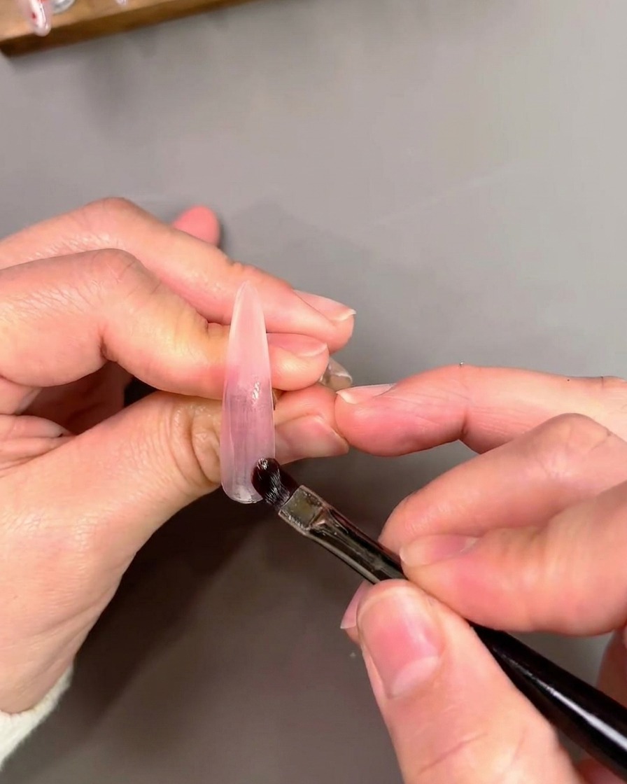 Un pennello piatto da gel che applica e sfuma lo smalto gel cat-eye viola su una tip artificiale.
