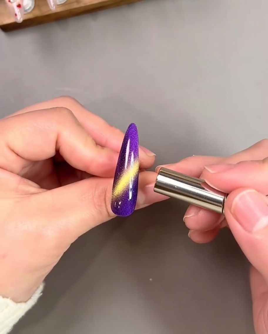 Una mano che tiene un magnete cilindrico vicino a ununghia dipinta di viola vibrante, attivando una striscia cat-eye oro brillante.