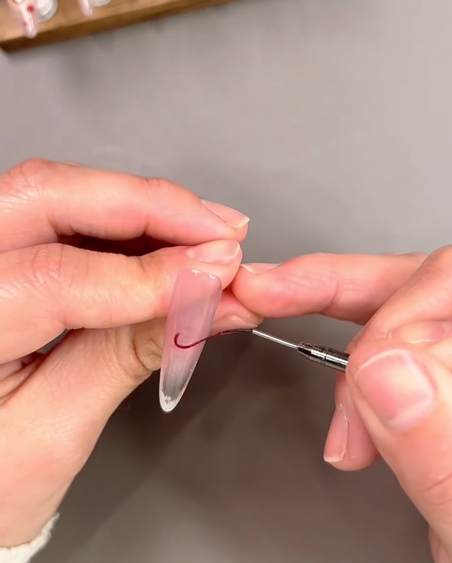 Un pennello a punta fine che disegna una sottile linea rossa curva in gel vicino al bordo libero di ununghia per contornare una punta French.