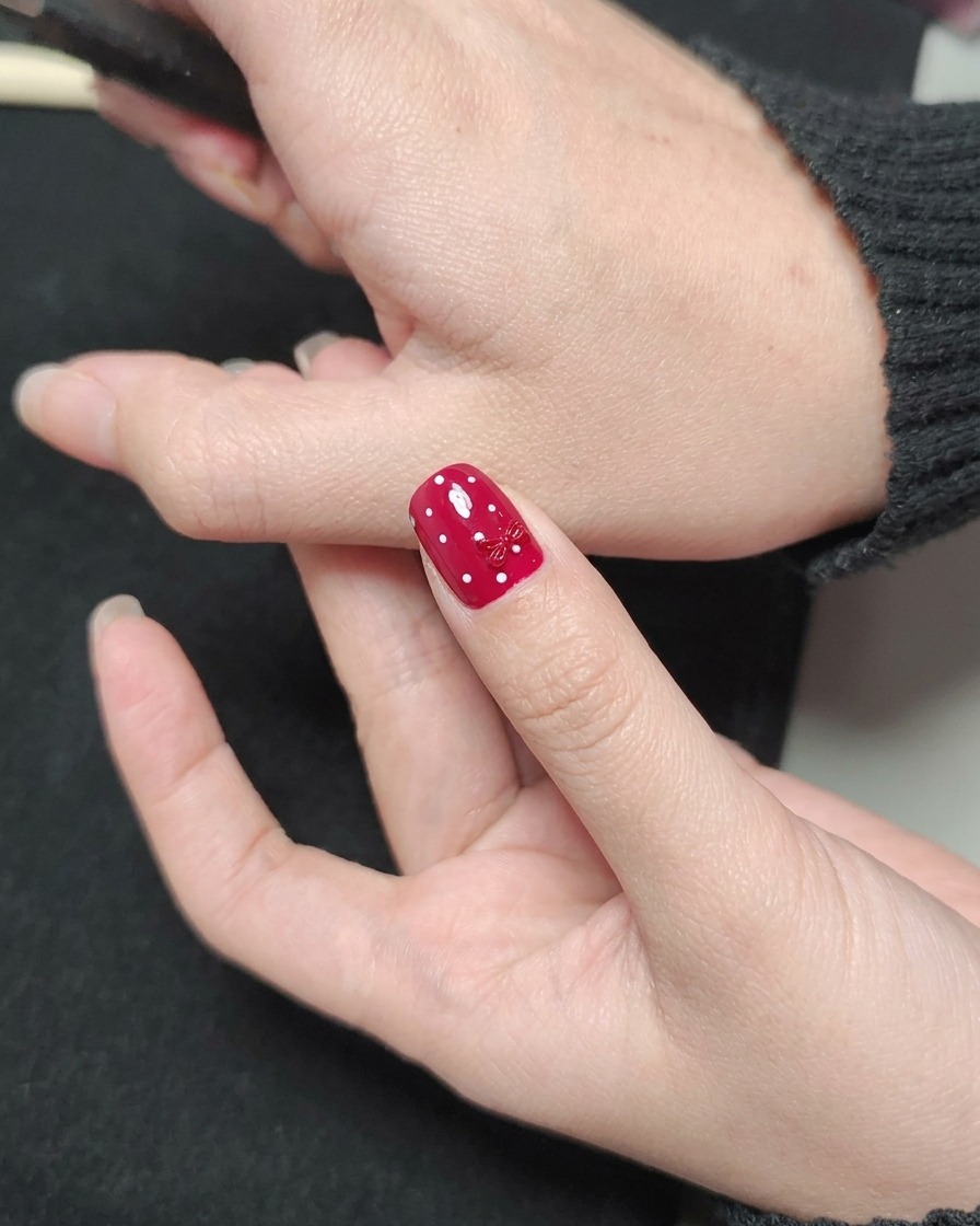 Un pennello per smalto applica un top coat trasparente e lucido su ununghia rossa a pois bianchi con un charm a forma di fiocco.