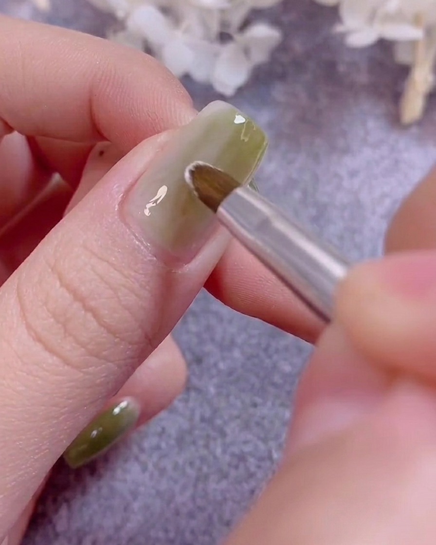 Un pennello sottile per nail art dipinge delicati petali di fiori bianchi su ununghia con base in gel verde.