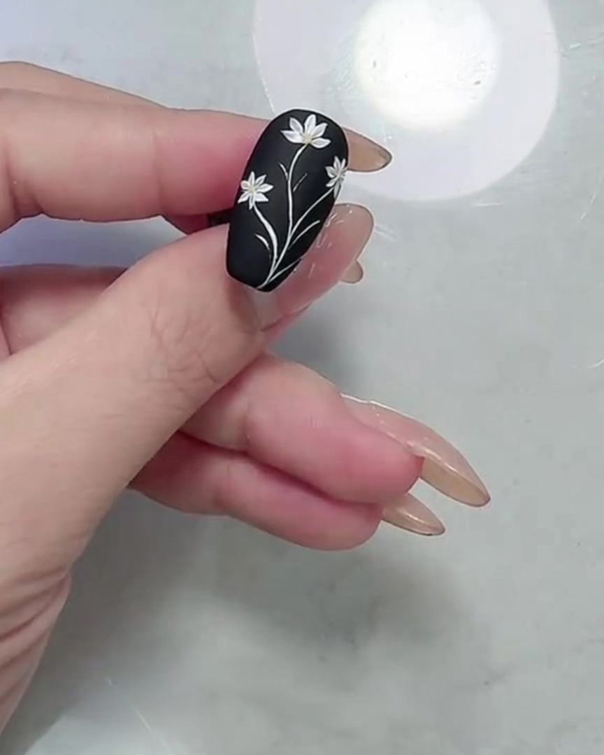 Uno scatto macro che mostra il design completo di margherite e foglie su ununghia nero opaco, concentrandosi sui bordi raffinati dei petali e delle foglie.