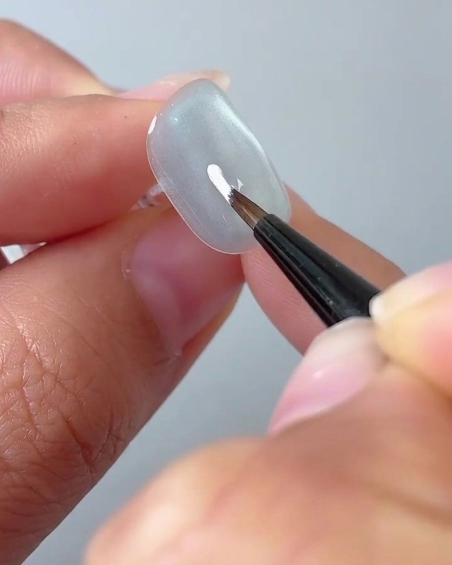 Un pennello liner fine che disegna un petalo bianco su una superficie ungueale grigio chiaro.