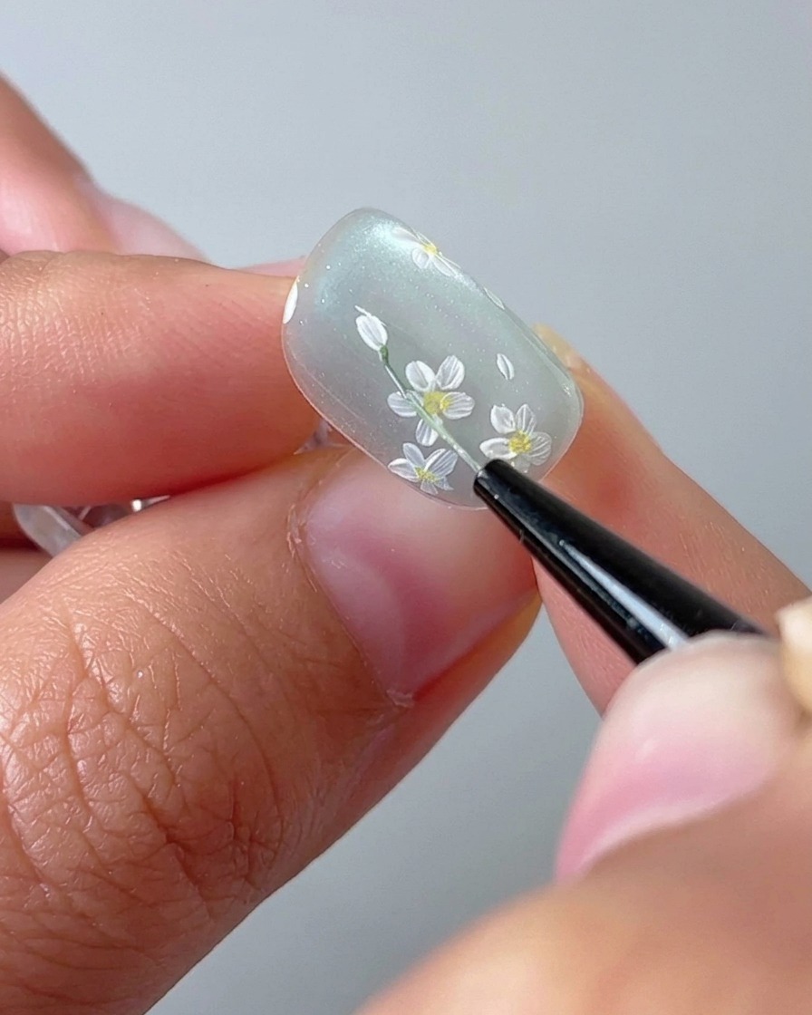 Un primo piano di un pennello che disegna un sottile gambo verde su una base gel cat-eye.