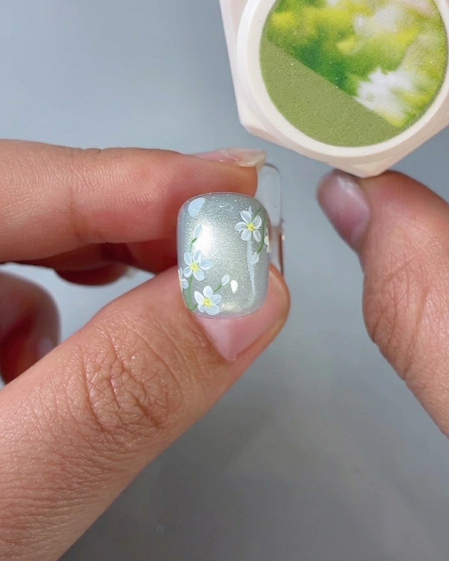 Un primo piano di un pennello che applica piccoli dettagli di foglie verdi su ununghia con arte floreale.