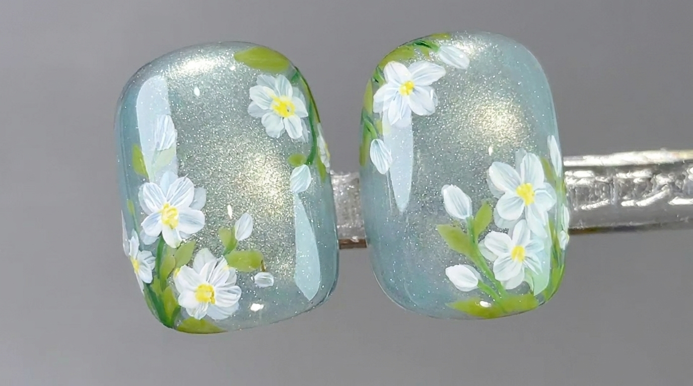 Un primo piano dettagliato di unghie in gel cat-eye trasparenti decorate con margherite bianche dipinte a mano e fogliame verde tenue.