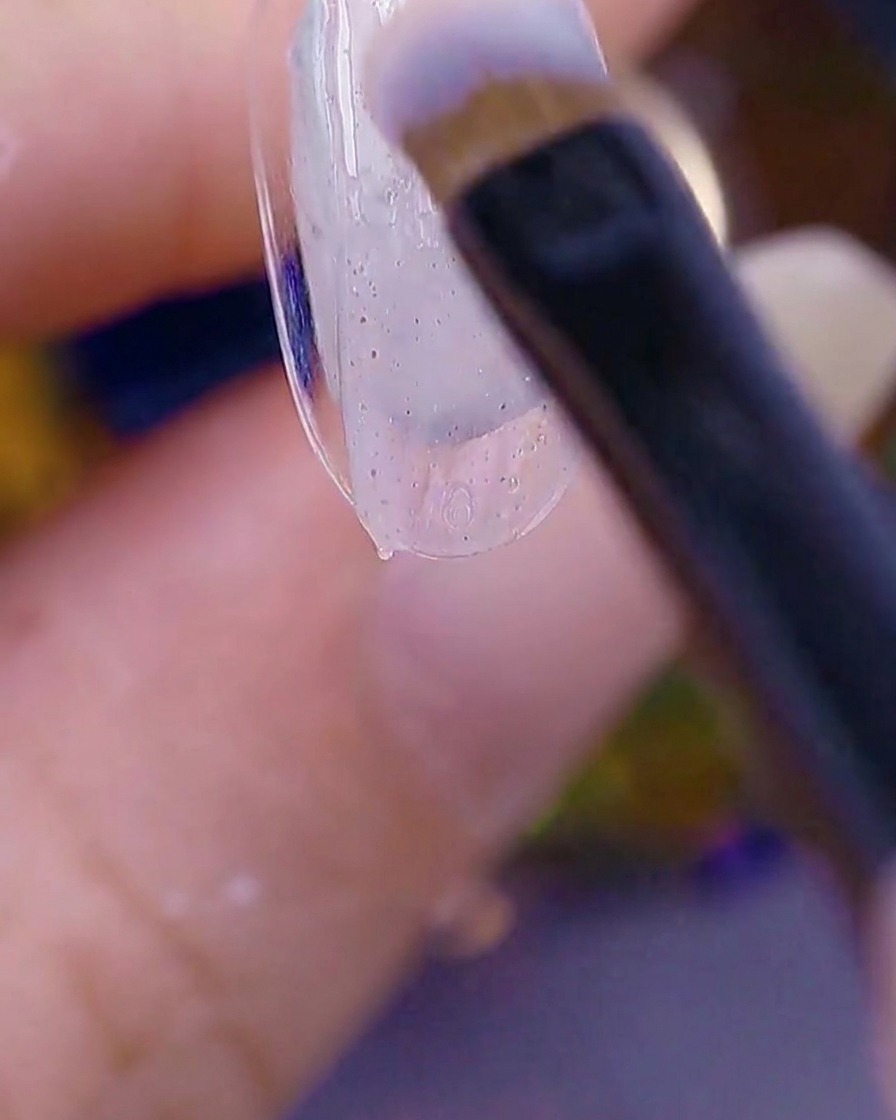 Primo piano di un pennello che applica un gel trasparente con texture di zucchero sulla punta di ununghia naturale.