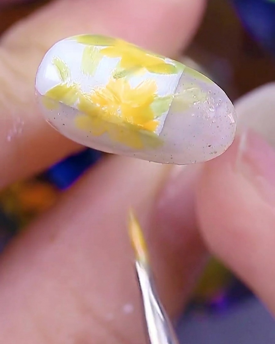 Primo piano dellunghia che mostra diversi fiori di margherita giallo-arancio dipinti sulle basi delle foglie, con il pennello che finisce gli ultimi petali.