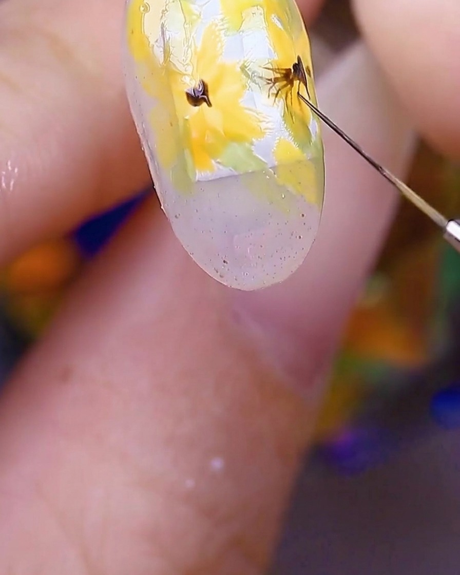 Primo piano della nail art che mostra un pennello che tira linee fini e sottili marrone scuro dal centro dei fiori gialli verso lesterno nei petali.