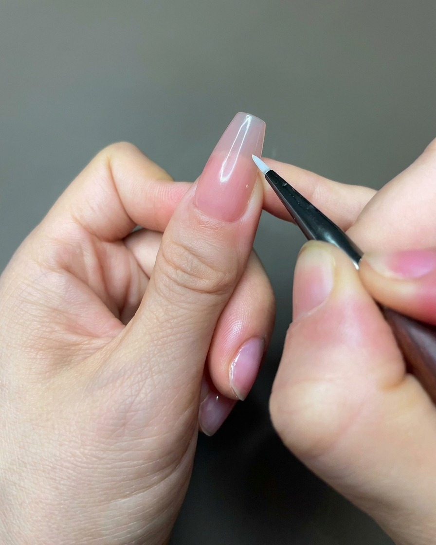 Un primo piano di un pennello liner fine che applica gel bianco per creare forme di petali su ununghia color nude.