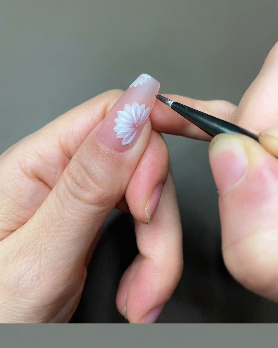Una vista ravvicinata di unonicotecnica che usa un pennello liner fine per aggiungere dettagli in gel dorato al centro di una margherita bianca su ununghia nude.