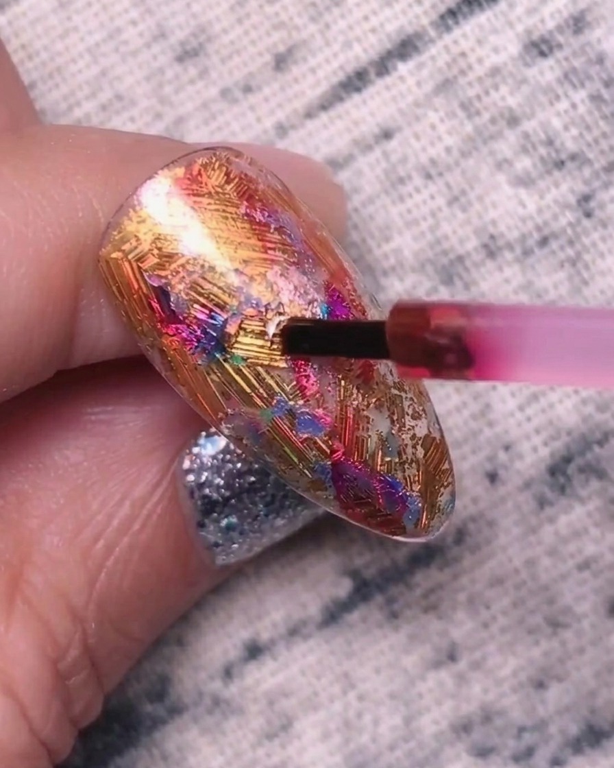 Un piccolo pennello applica un gel pigmentato rosso-viola su una superficie ungueale testurizzata coperta di foil metallizzati.