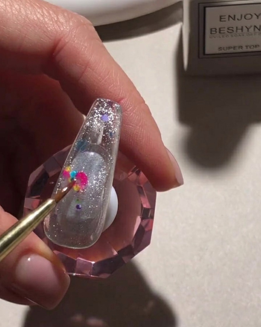 Un pennello di precisione posiziona vivaci paillettes rotonde multicolori su una superficie ungueale glitterata.