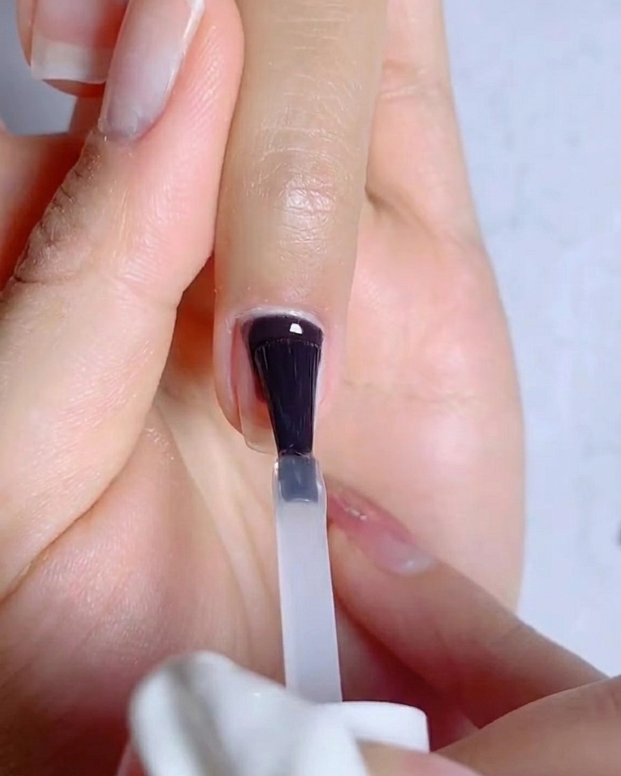 Un primo piano di un pennello che applica uno strato sottile e trasparente di base coat gel su ununghia naturale preparata.