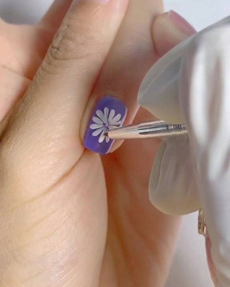 Un dito con la nail art della margherita bianca completata su una base viola posizionato sotto una piccola lampada professionale per unghie per la polimerizzazione.