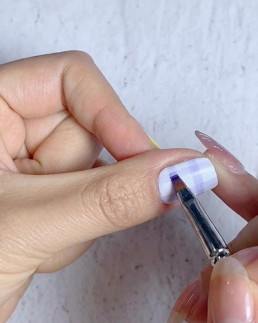 Un pennello sottile che riempie le intersezioni delle linee della griglia tartan su un design di unghia con gel blu.