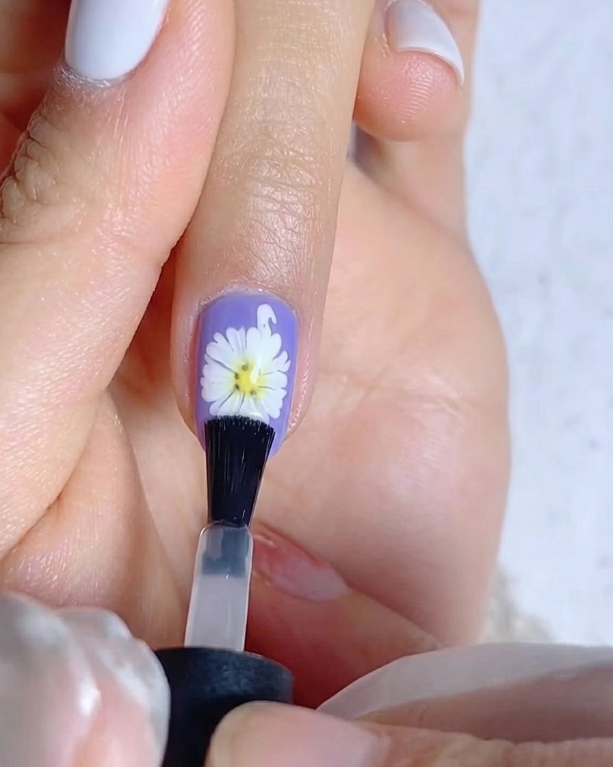 Un pennello che applica uno strato di gel top coat opaco su ununghia viola caratterizzata da un design a margherita bianca.