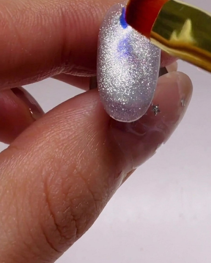 Un piccolo pennello posiziona glitter iridescenti blu e viola su ununghia argentata.