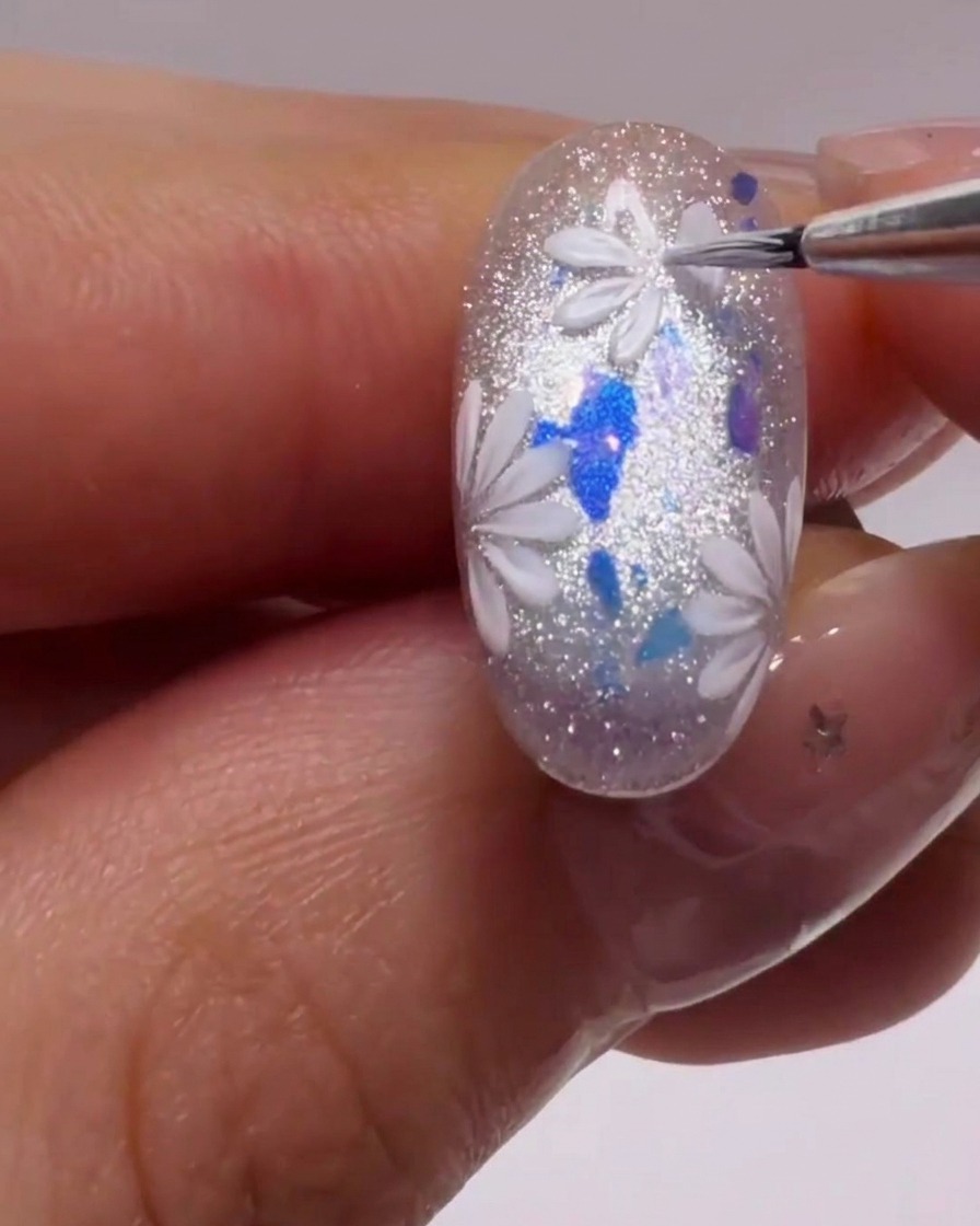 Un pennello liner fine crea petali bianchi precisi su ununghia glitterata.