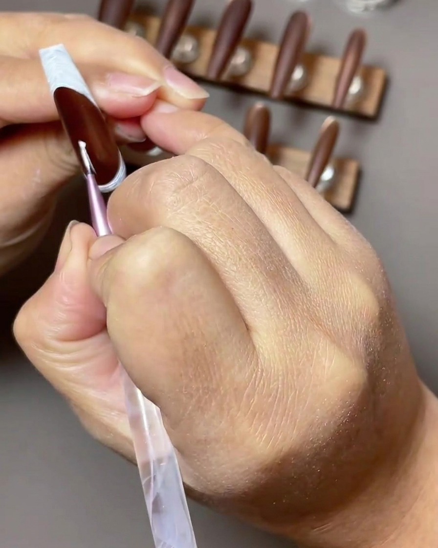 Una vista ravvicinata di un pennello sottile da nail art che applica una stella bianca a quattro punte in gel su ununghia marrone scuro con una french tip inversa.