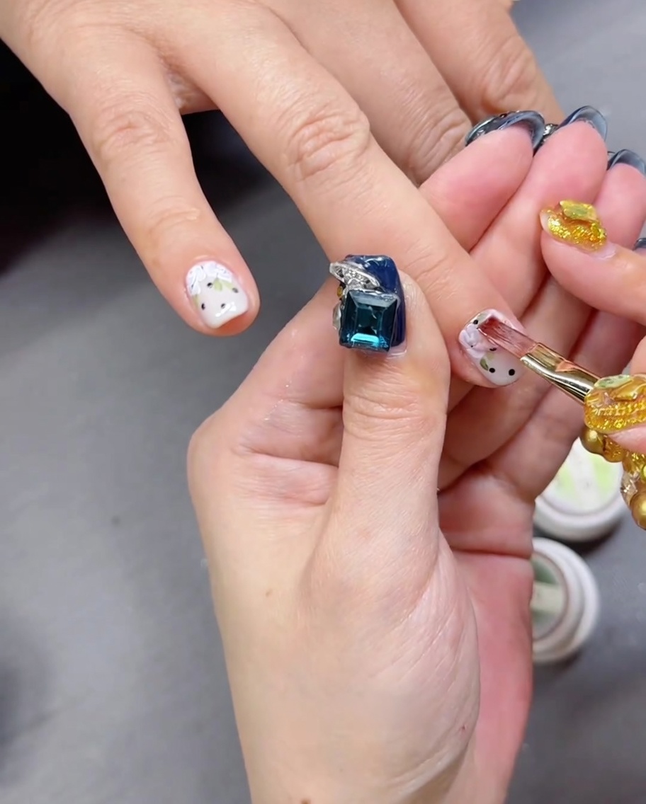 Primo piano di un pennello che applica un top coat trasparente e lucido sopra ununghia che presenta un design di rose dipinte a mano e pois.