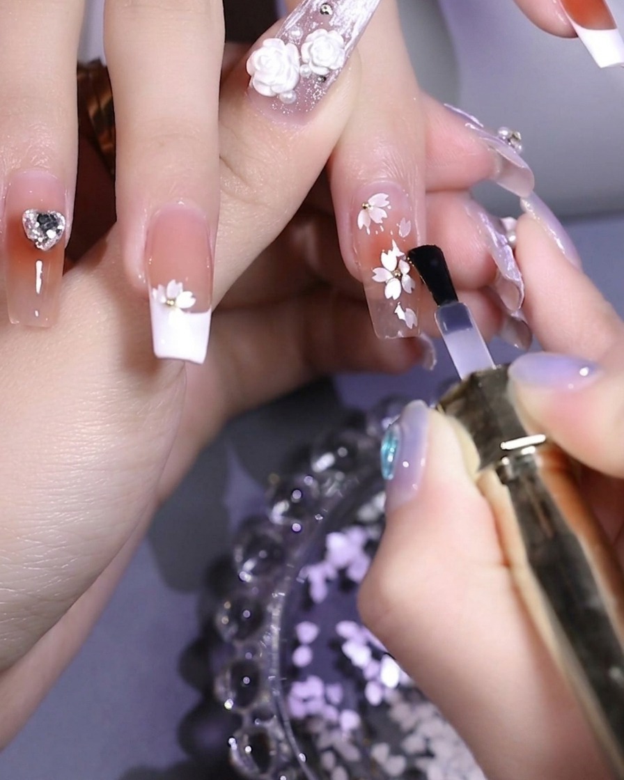 Un pennello che applica un top coat lucido trasparente su ununghia decorata con pailettes floreali bianche.