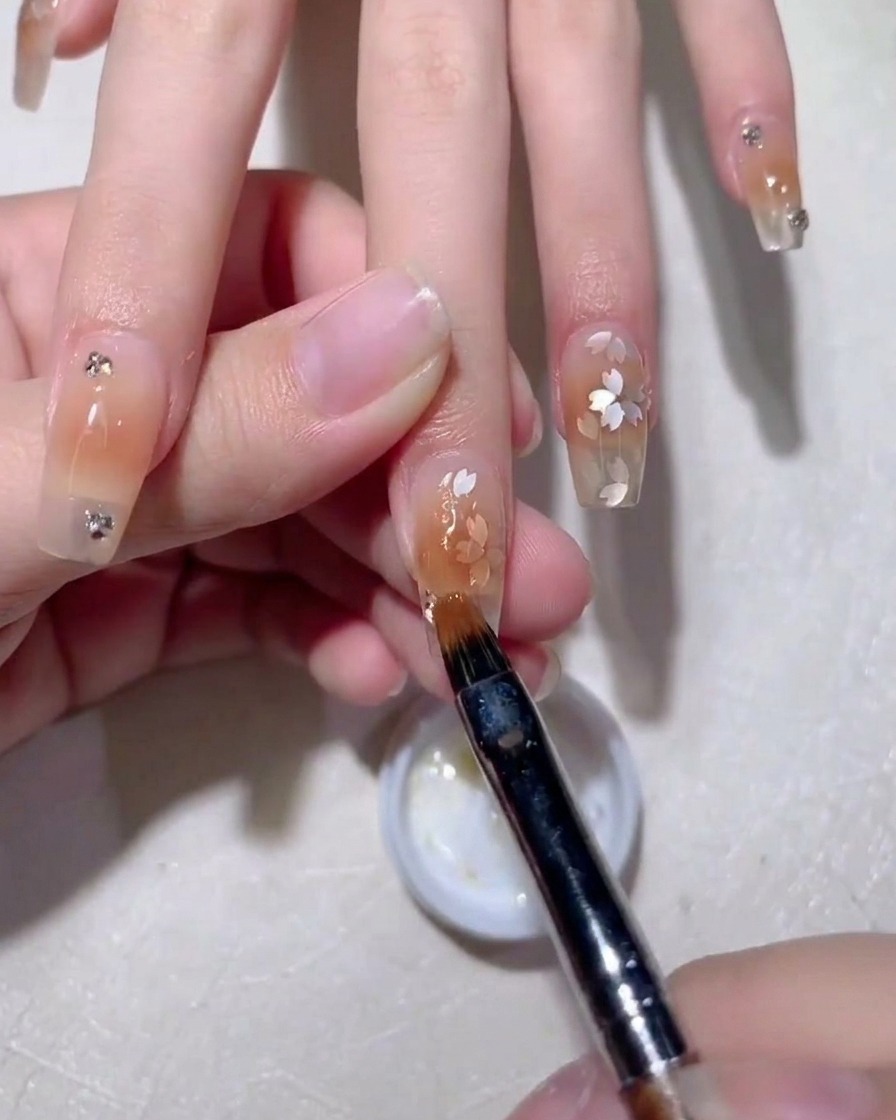 Una vista ravvicinata di un pennello per nail art che liscia il gel costruttore trasparente sopra delicate paillettes a forma di fiore di ciliegio su ununghia color nudo.