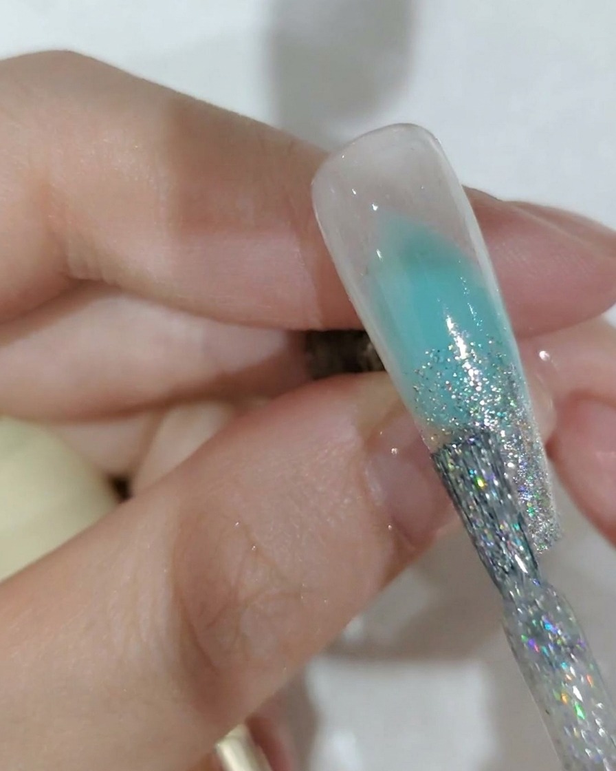 Un primo piano di un pennello da unghie che applica gel glitter argento sulla punta di ununghia.