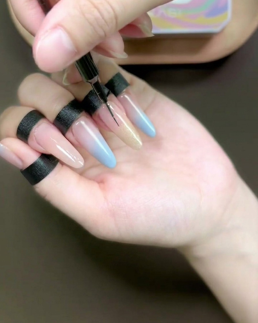 Un primo piano di un pennellino fine che sfuma il gel cat-eye color champagne su un anulare lungo con ricostruzione.