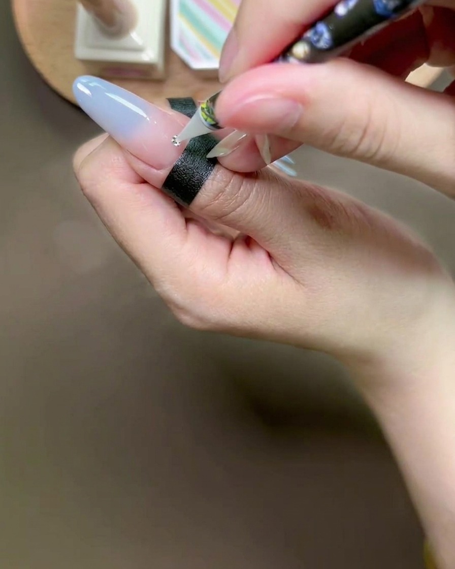 Una penna in cera posiziona un piccolo strass argentato su una goccia di colla su ununghia.