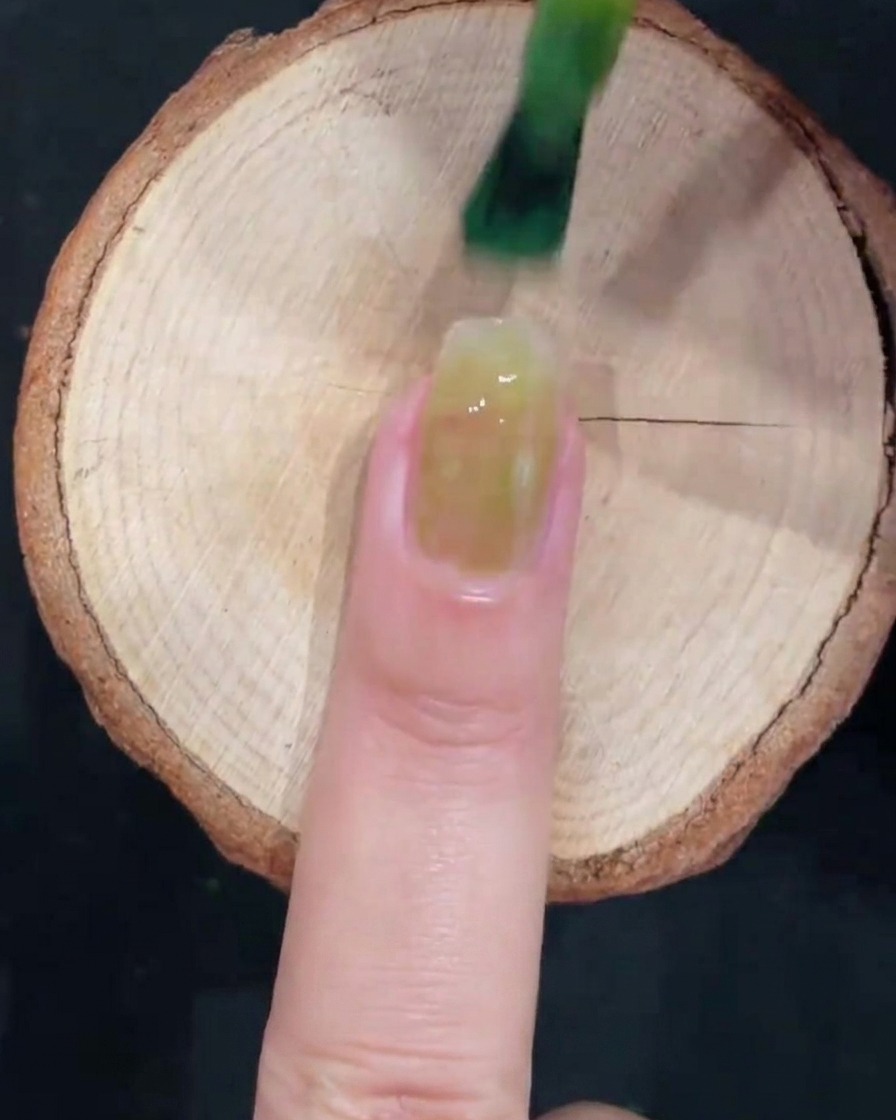 Un pennello da gel applica uno smalto verde ghiaccio traslucido al centro di ununghia naturale pulita.