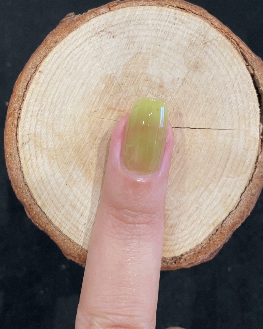 Dettaglio di un pennello che applica gel verde traslucido su ununghia naturale.