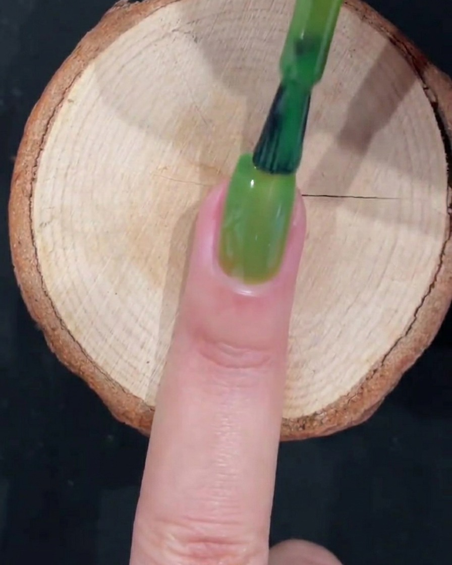 Un pennello da gel completa uno strato di smalto verde traslucido con pennellate fluide lungo lunghia.