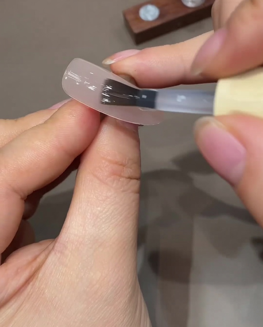 Un pennello piatto per gel che applica uno strato sottile e trasparente di top coat su ununghia preparata color nude.