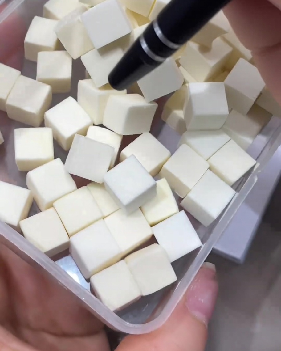 Una pinzetta di precisione che preleva un piccolo cubetto di spugnetta per trucco bianca da un contenitore di plastica trasparente.