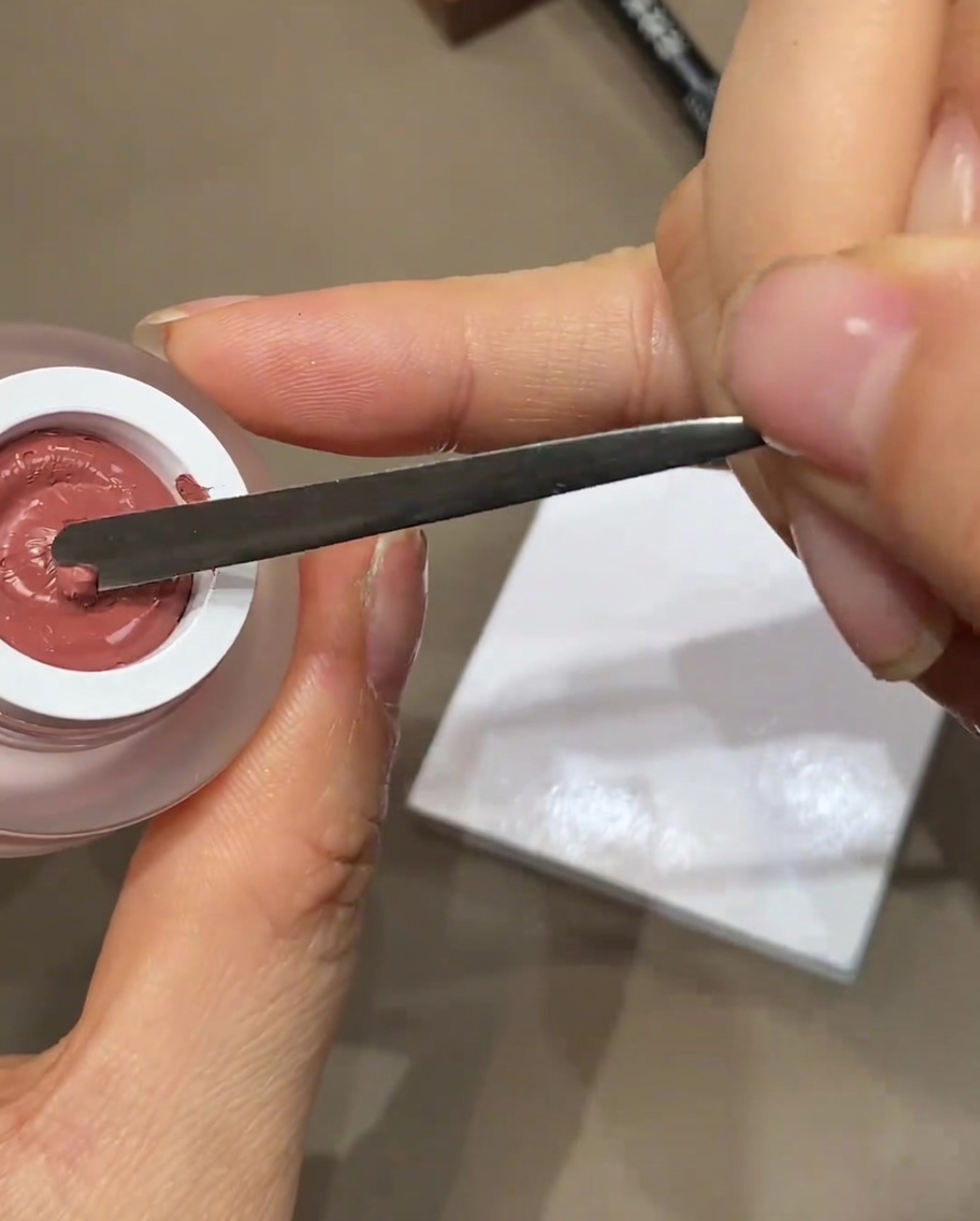 Una spatola in metallo che versa una piccola goccia di smalto gel colorato su una tavolozza bianca.