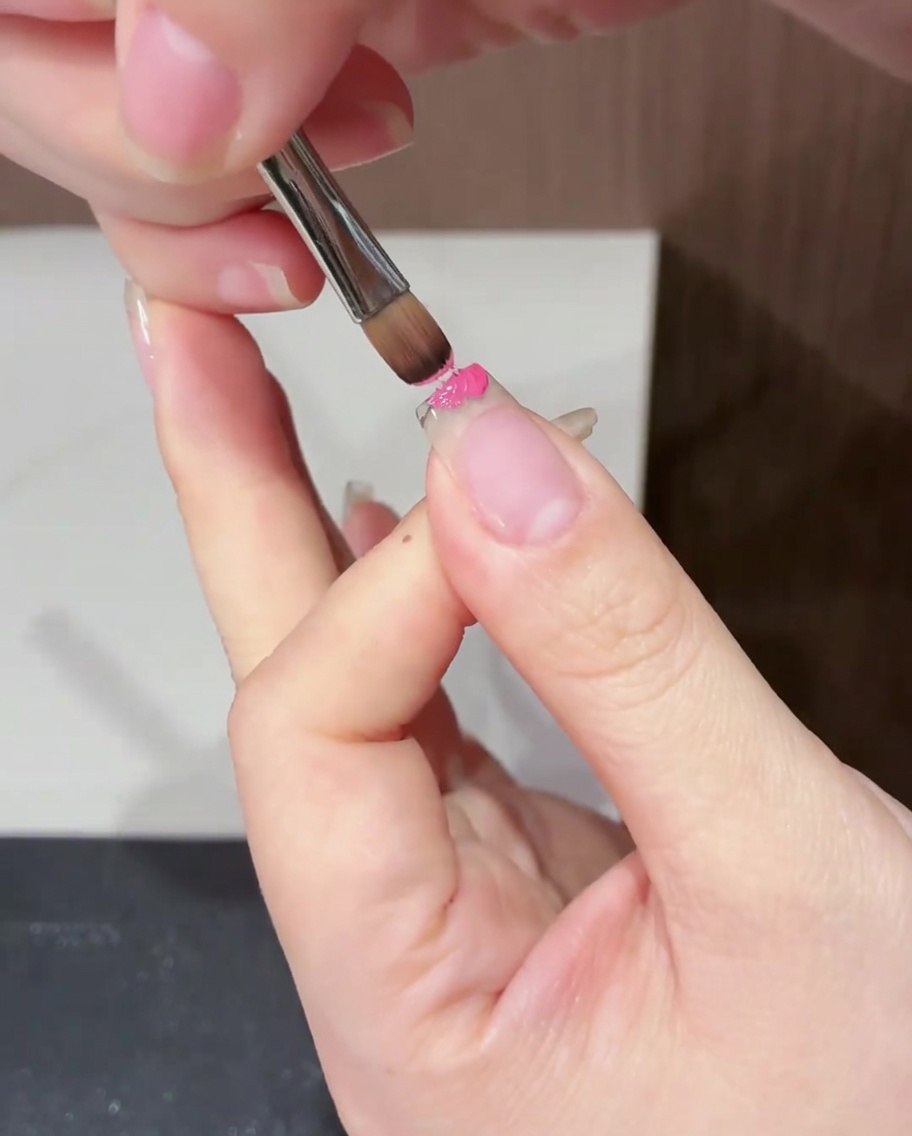 Un primo piano di un pennello piatto che applica smalto gel magenta specificamente sul margine libero di ununghia naturale.