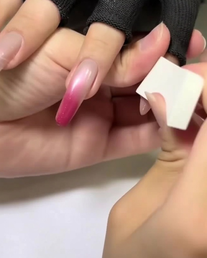 Una mano che utilizza una spugnetta per makeup bianca per applicare un colore rosa chiaro sulla parte superiore di ununghia a mandorla.