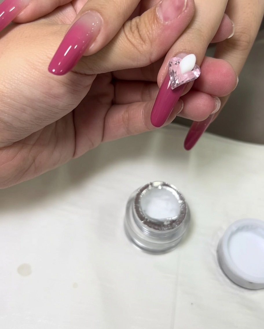 Una goccia di gel da scultura 3D bianco posta sullunghia accanto a un cristallo a cuore rosa.
