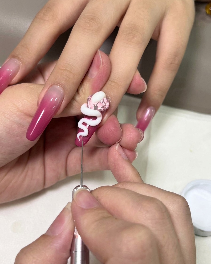 Un primo piano di una mano che usa uno strumento di precisione in metallo per modellare un serpente in gel 3D bianco sopra un cristallo rosa su ununghia ombré magenta.