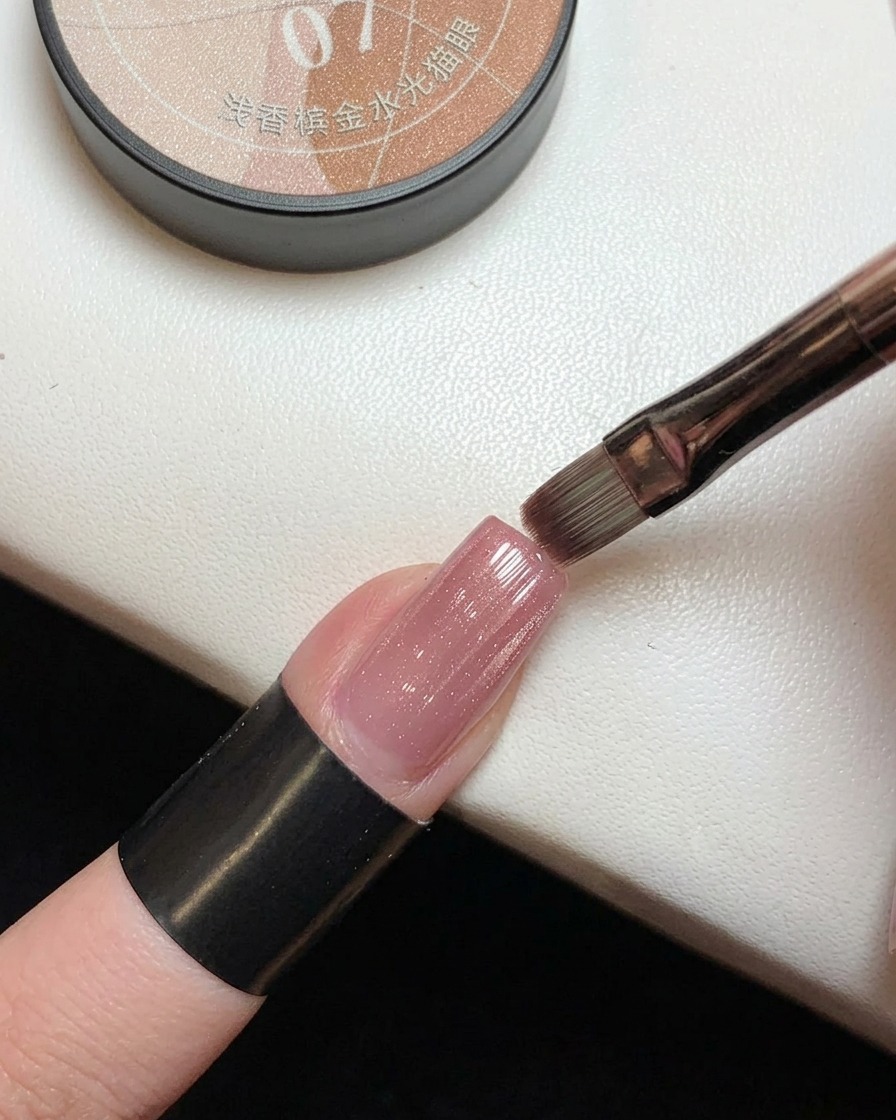 Un pennello che applica un gel magnetico rosa traslucido con riflessi dorati sopra ununghia con sfumatura rosa già polimerizzata.