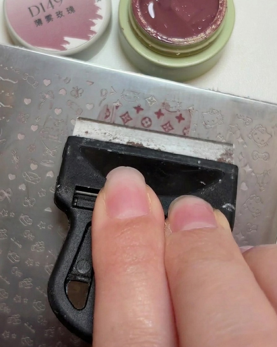 Un raschietto di plastica che livella una piccola noce di smalto gel rosa antico attraverso i motivi incisi su una piastra metallica per stamping.