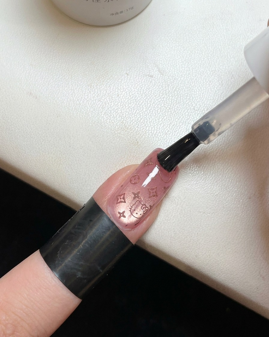 Un pennello che applica un top coat trasparente e lucido su ununghia decorata con un motivo rosa di Hello Kitty.