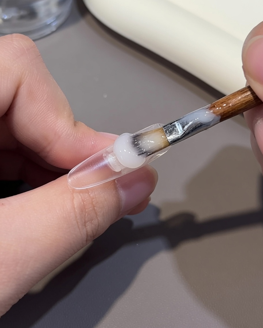Un pennello applica un gel color nude sulla parte basale di una tip unghia trasparente.