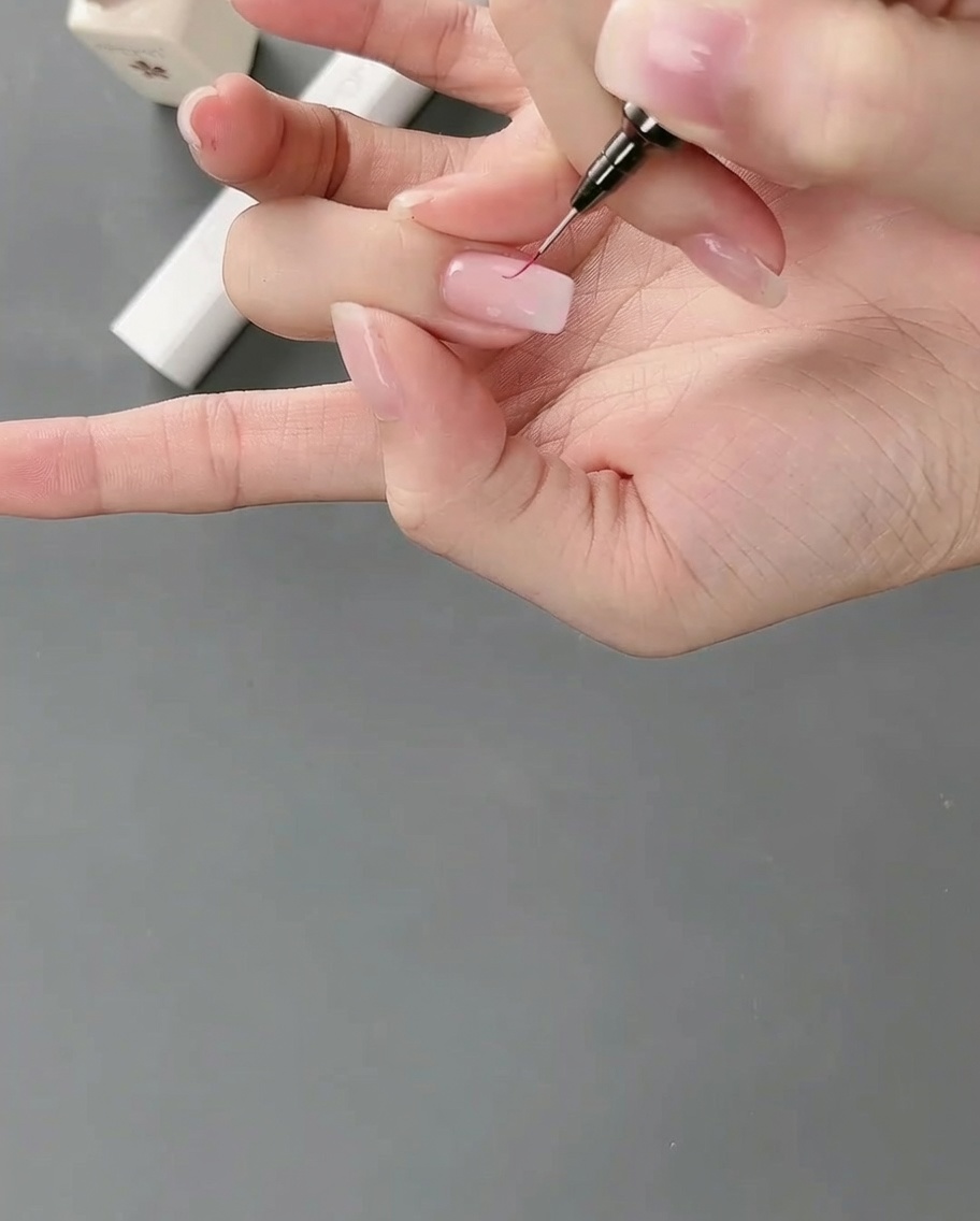 Primo piano di un pennello liner che disegna linee sottili e dettagliate attorno al bordo di un blocco di gel rosa su ununghia.