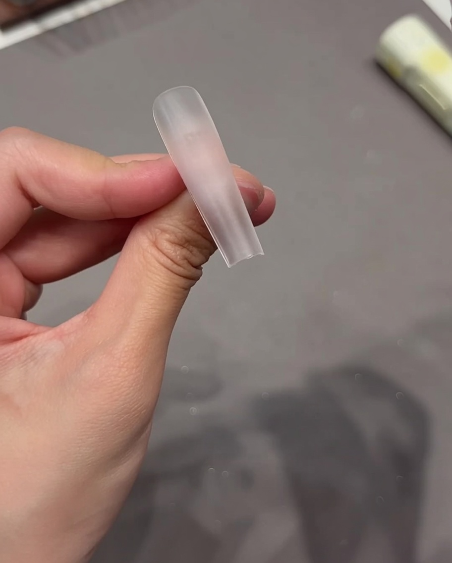 Primo piano di una mano che applica una linguetta adesiva trasparente sulla superficie di una tip per unghie chiara.