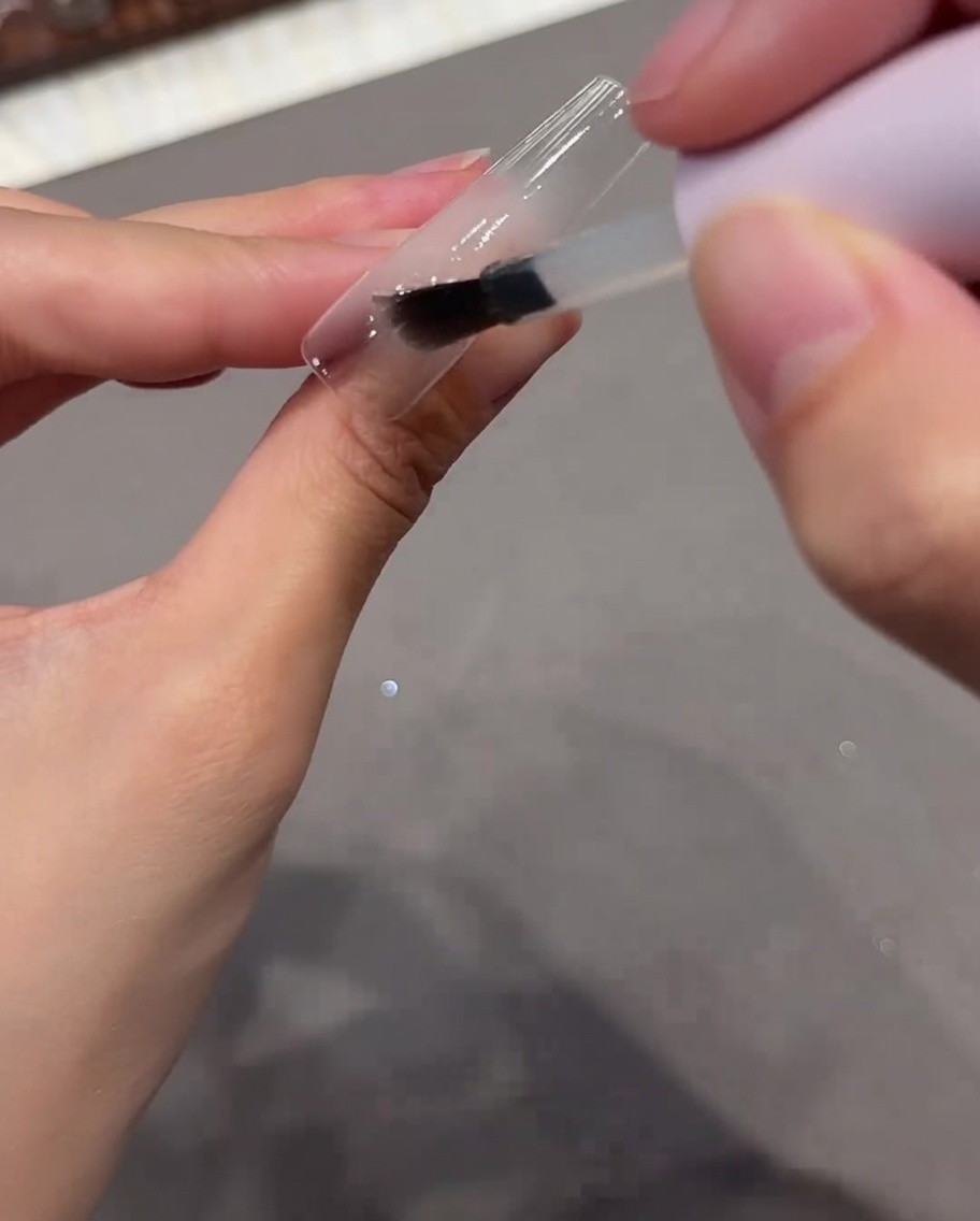 Un pennello che applica una base coat trasparente su una tip per unghie chiara.