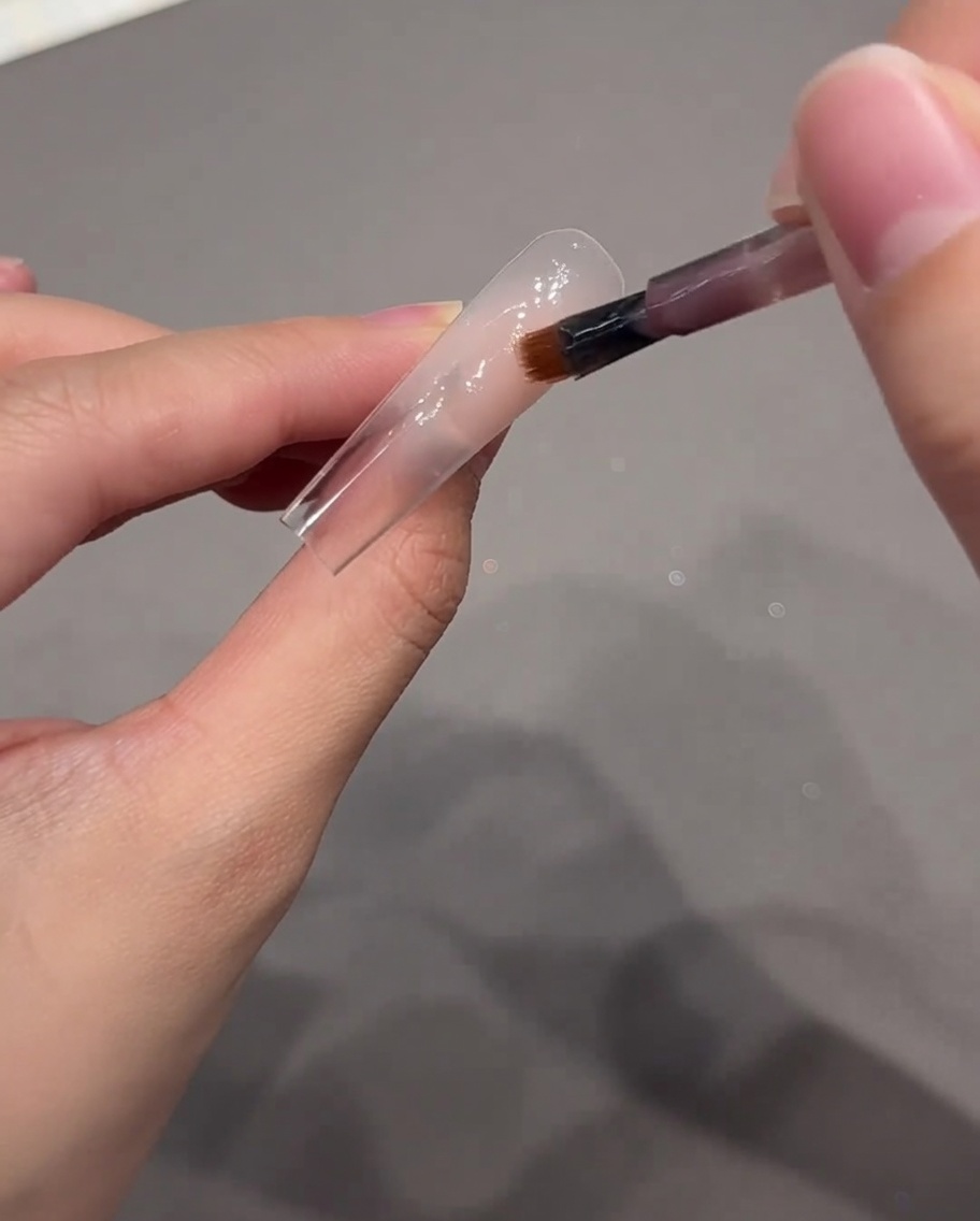 Un pennello per nail art che applica una goccia di gel rosa nude sulla zona delle cuticole della tip.